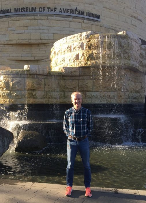 Harrison stands in front of the water featuer at NMAI, wearing jeans and pink sneakers