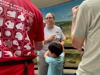 Jen Ferraro, wearing gray staff T-shirt, is seen between the backs of visitors listening as she speaks