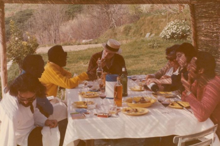 Color photo of James Baldwin and several unidentified people sitting at an outdoor table with food and drink