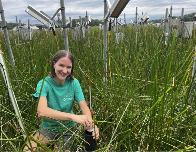 Genevieve Noyce in green T-shirt and shorts sits in a grassy field surrounded by heat lamps