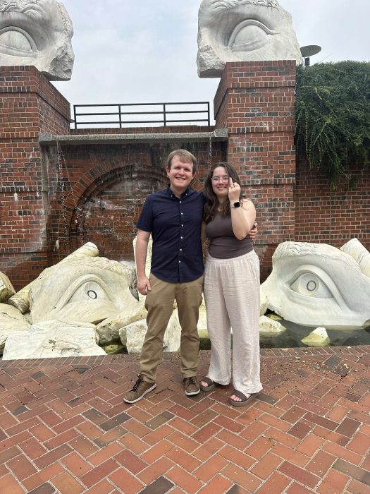 Couple stand before brick wall with sculptural pieces (Haines Point?) She is lifting one finger to show engagement ring.