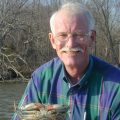 Tuck Hines, wearing a blue plaid shirt, holds a blue crab.