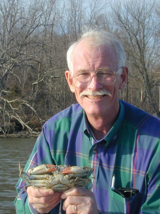 Tuck Hines, wearing a blue plaid shirt, holds a blue crab. 