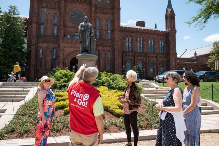 A volunteer wearing red vest talks to tourists on the Mall in front of the Smithsonian Castle