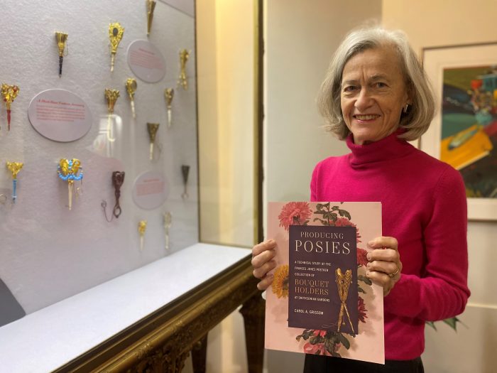 Carol Grissom, in pink sweater, holds a copy of her latest book while standing next to a display case of antique bouquet holders.