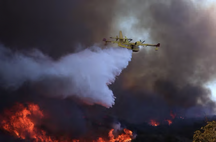 A plane drops water on wildfire against backdrop of smoky sky