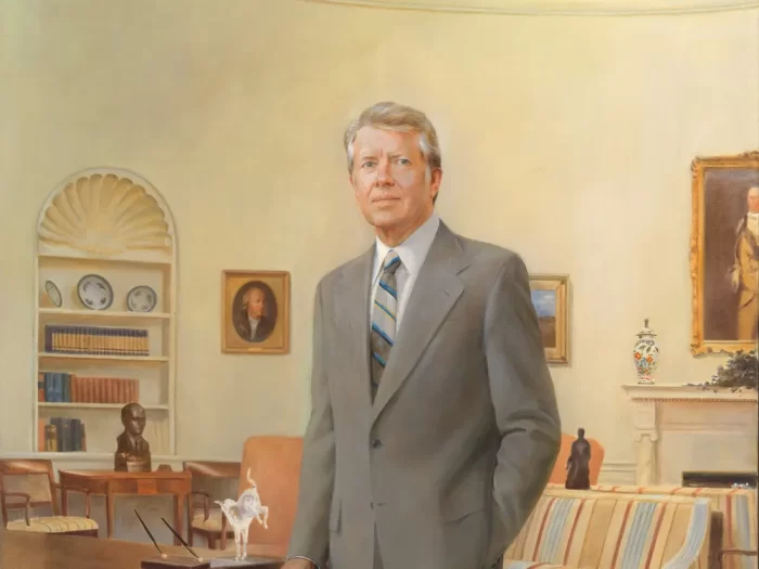 Jimmy Carter stands next to his desk in the Oval Office, dressed in a gray suit