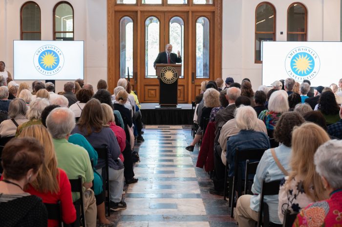 Bunch at dais with screens showing Smithsonian logo to his right and left